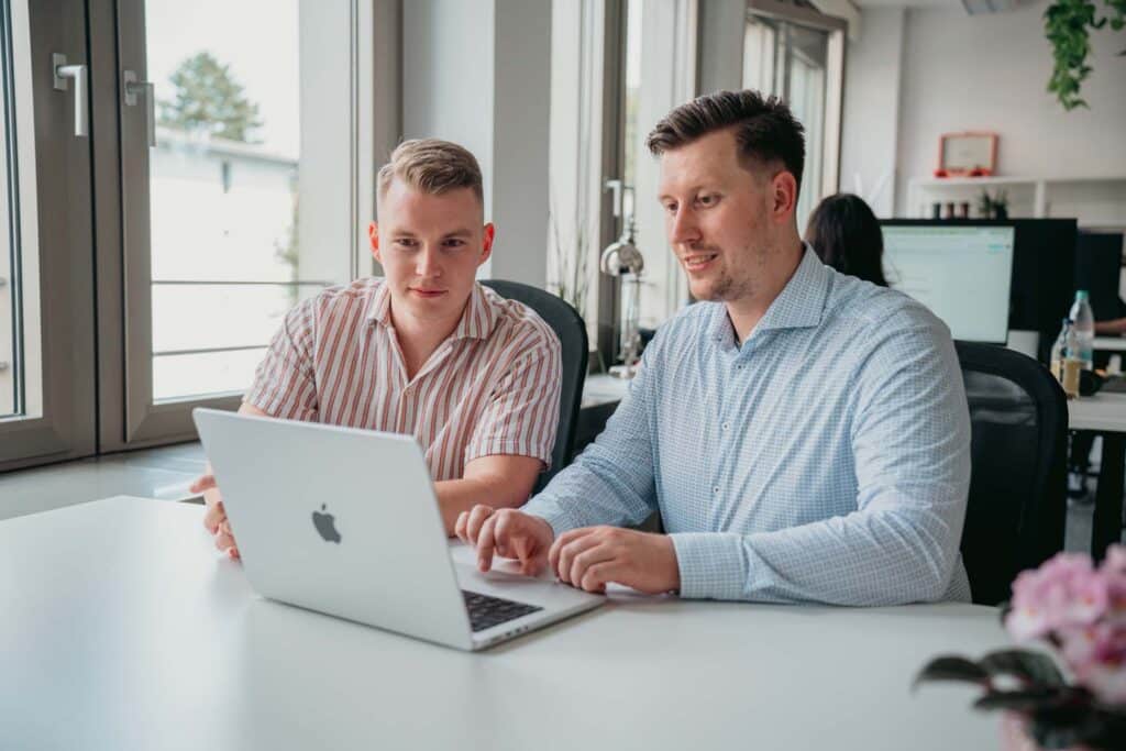 Zwei Männer sitzen an einem Schreibtisch in einem modernen Büro und schauen gemeinsam auf einen Laptop-Bildschirm. Konzentriert und engagiert arbeiten sie gemeinsam an der Digitalisierung des Einzelhandels. Große Fenster hinter ihnen lassen Tageslicht herein.